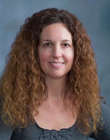 Woman with curly brown hair smiling, wearing a gray top, with a blue background.
