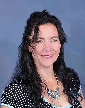 Woman with dark wavy hair smiles, wearing a polka dot top and a silver necklace, against a blue background.