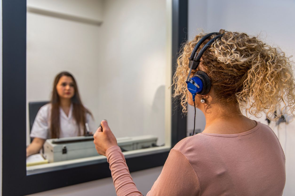 Woman in headphones having a hearing test. A professional looks on from the other side of the mirror.