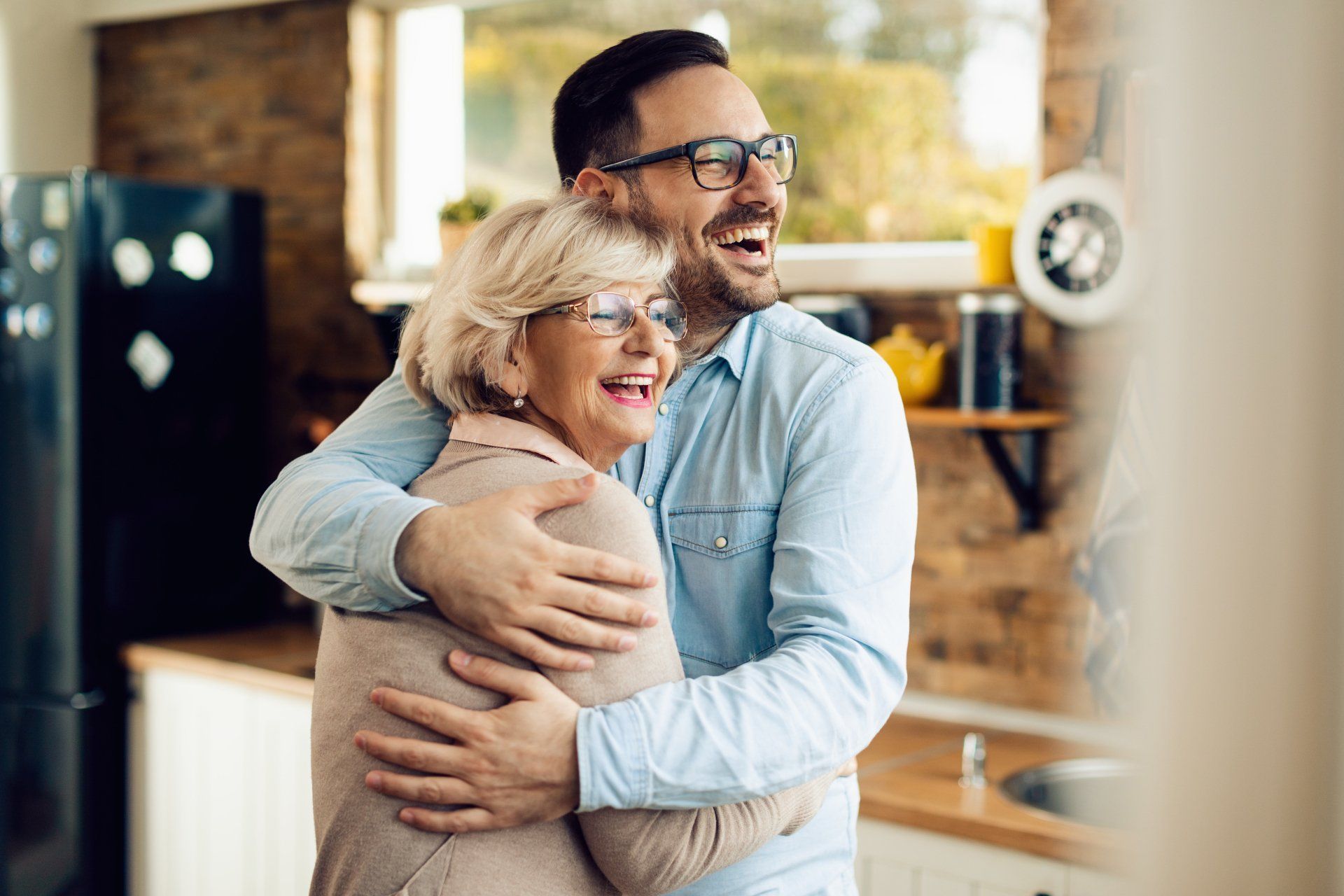 A man hugging an older person in a kitchen; both smiling, cheerful.