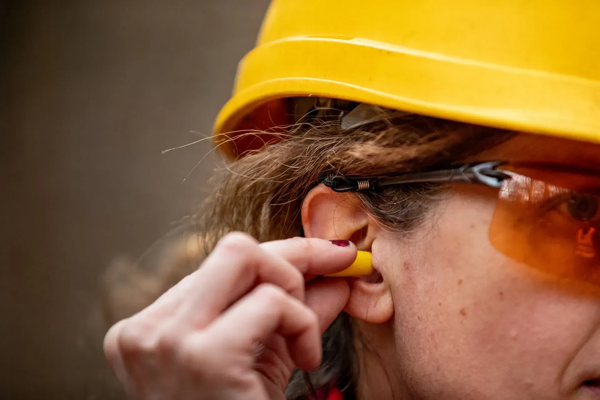 Person inserting a yellow earplug into ear while wearing a yellow hard hat and safety glasses.