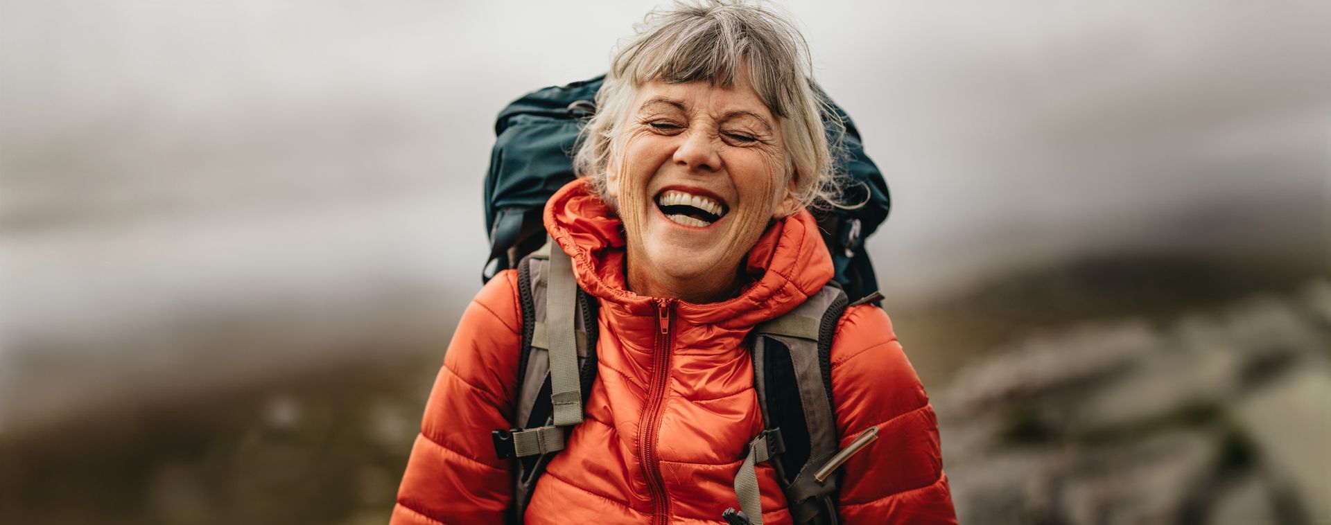 Woman with a backpack laughing while hiking in a blurred outdoor setting; she wears a red jacket.