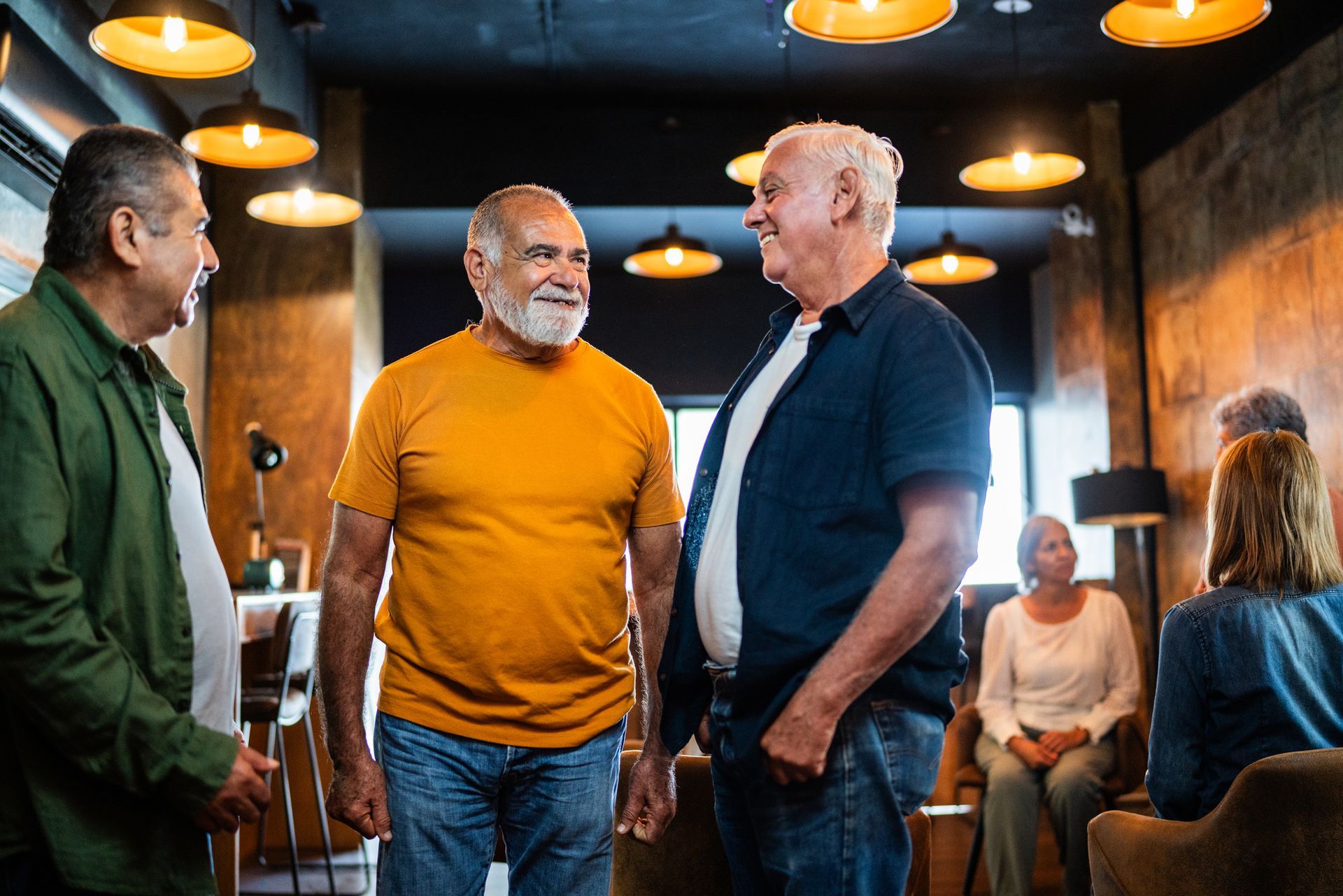 Three men conversing in a dimly lit room, with others seated in the background.