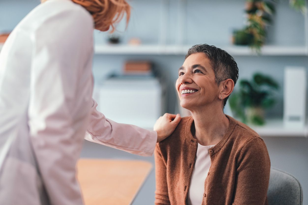 Woman smiles while a person in a white coat puts their hand on her shoulder in an office setting.