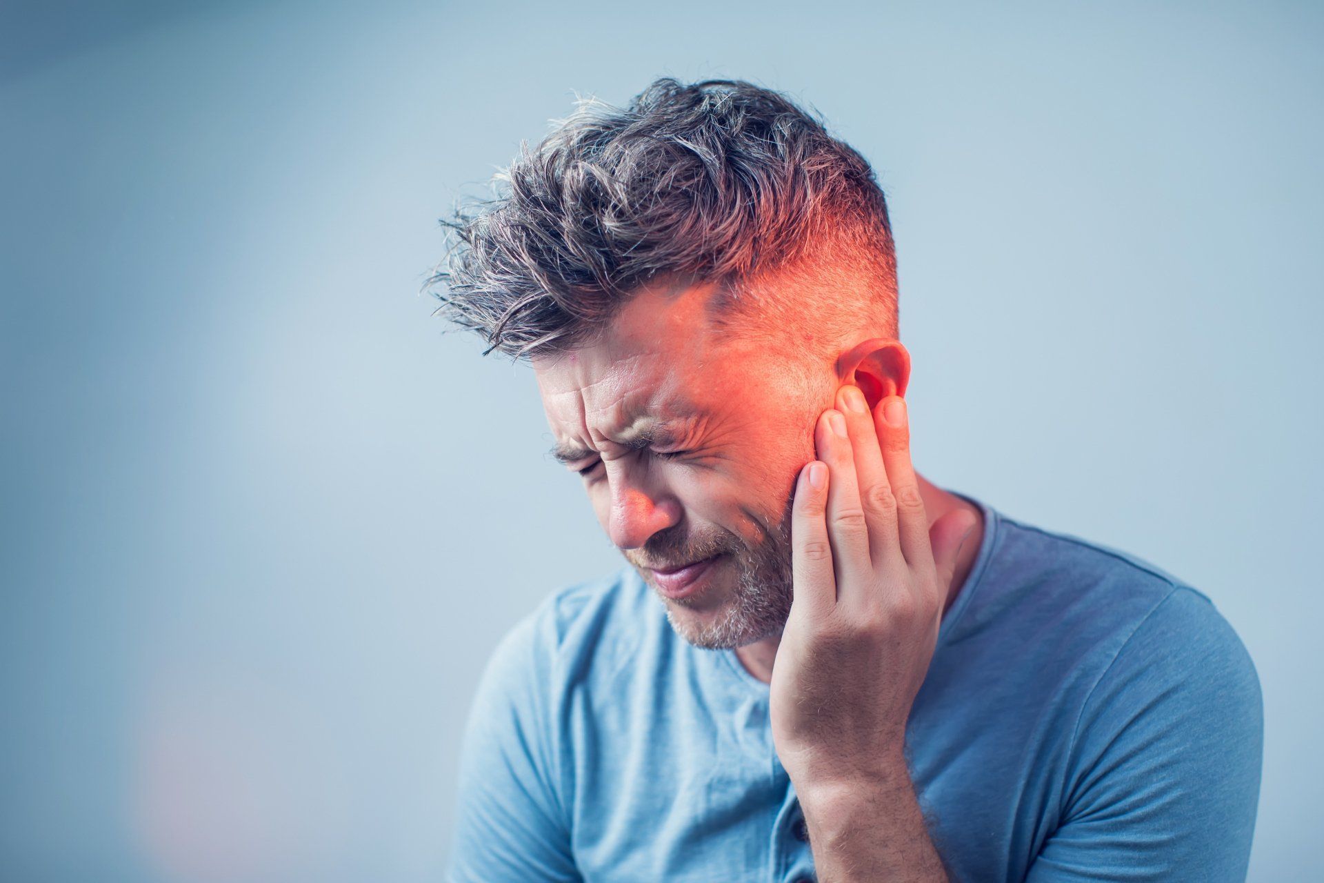 Man holding his ear, face in pain, red glow on face, light blue background.