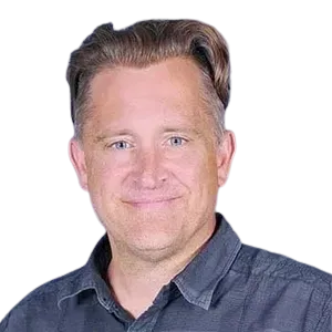 Man in a dark button-down shirt smiles, looking at the camera. He has fair skin and light brown hair.