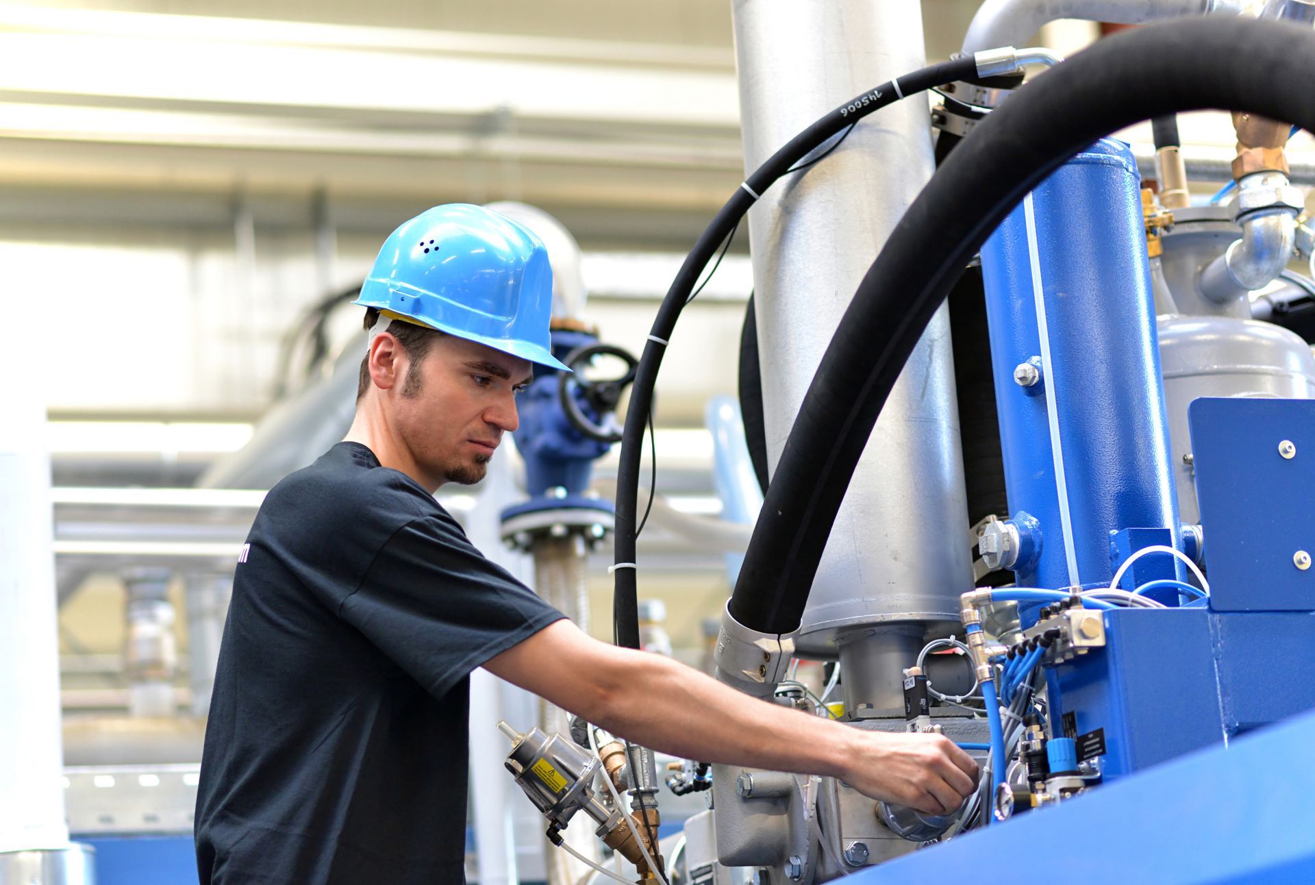A worker in a blue hard hat inspects machinery in a factory, touching a control panel.