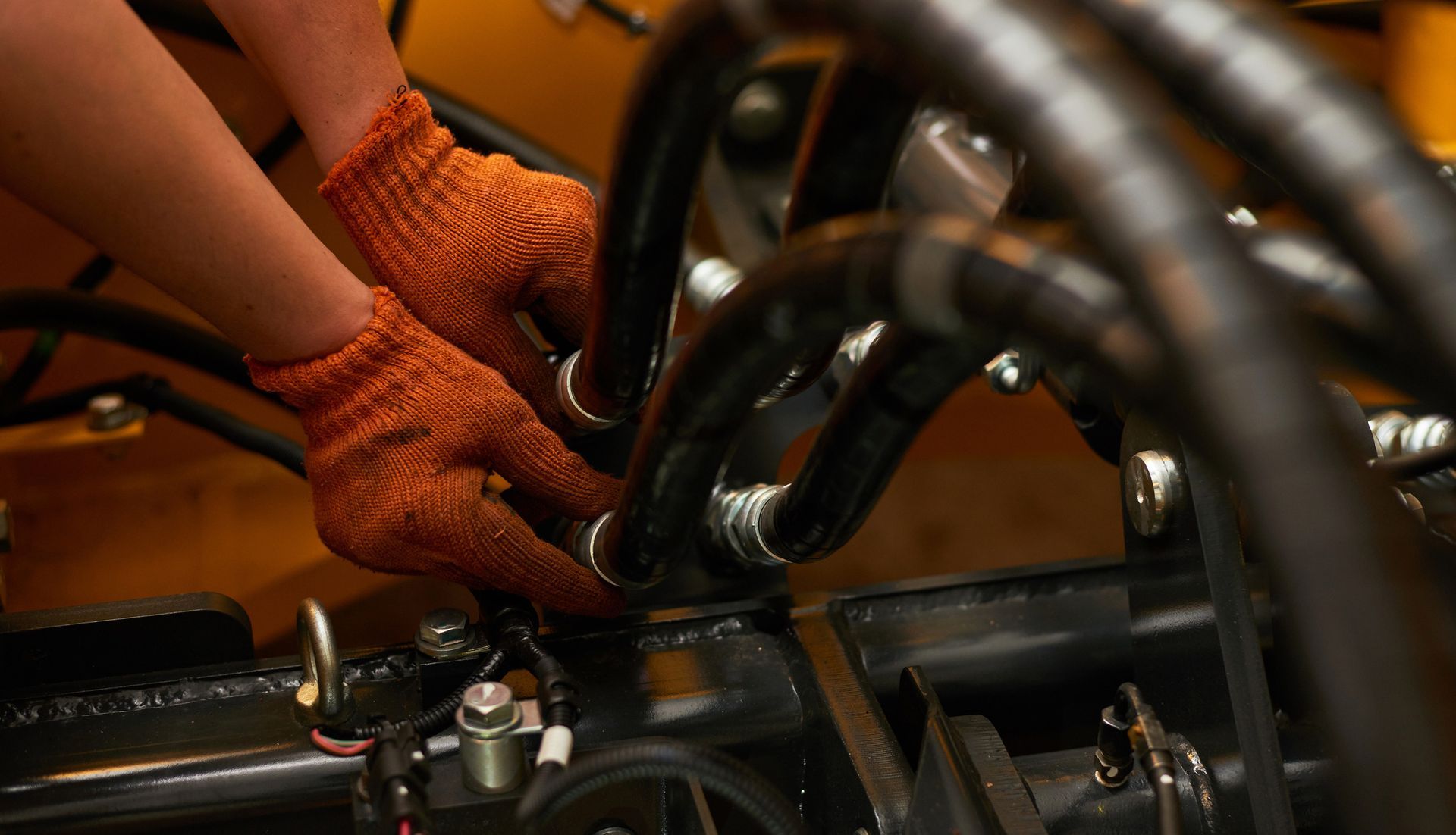 Hands in orange gloves working on black machinery with hydraulic hoses.