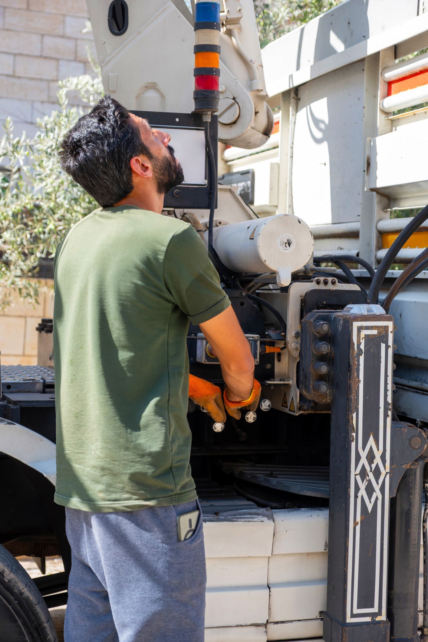 Man operating machinery, looking up. Wearing green shirt, grey pants. Orange lights. Outdoors.