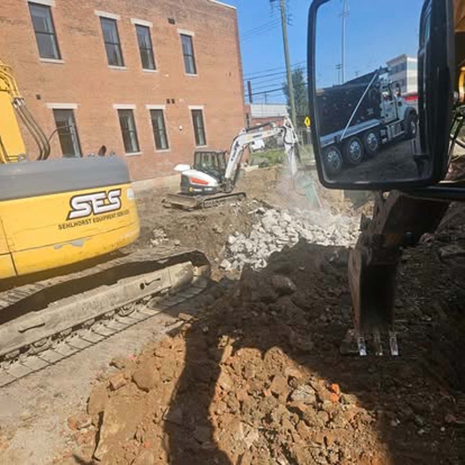 A yellow excavator is digging a hole in the dirt in front of a brick building.