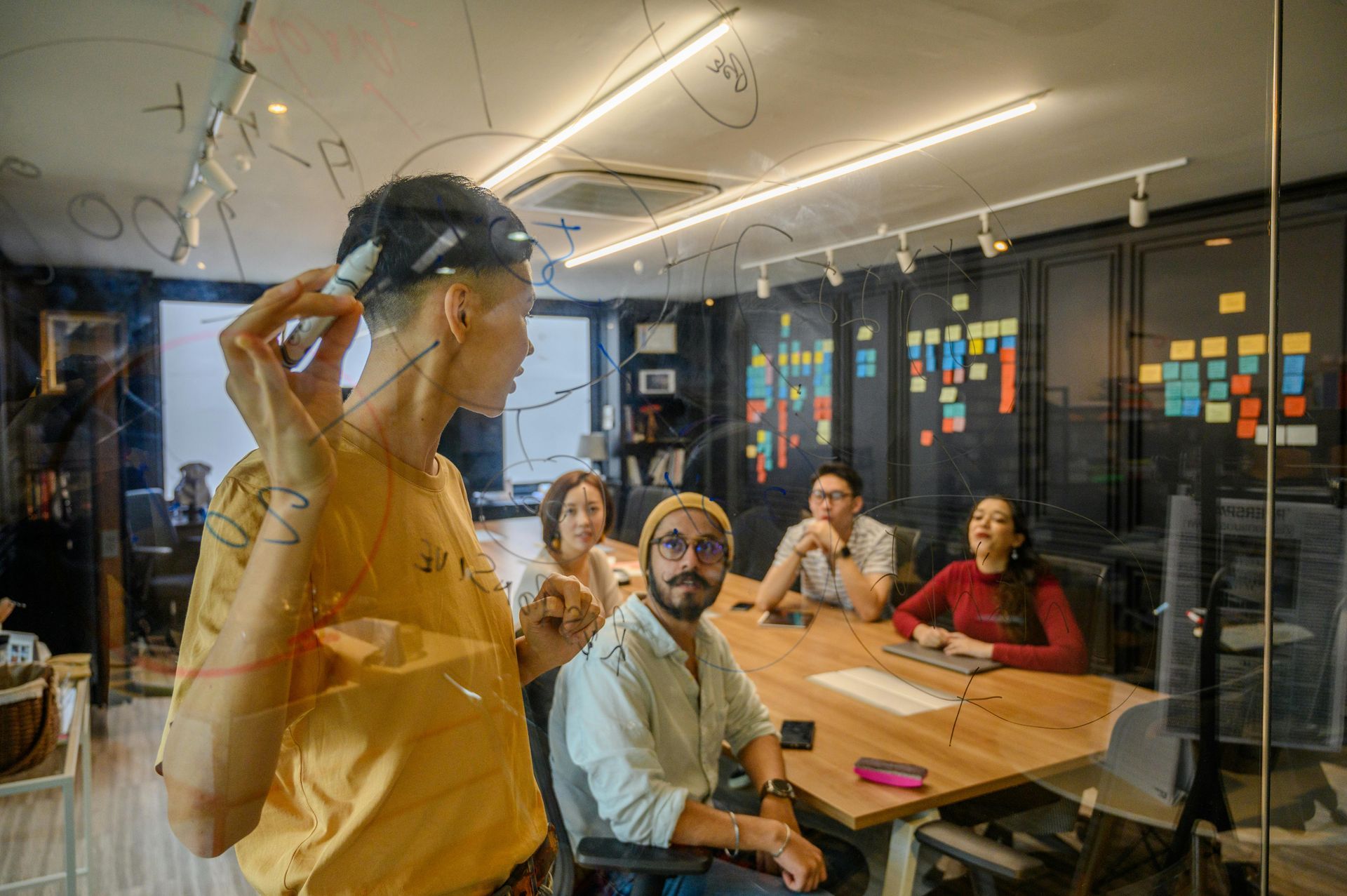 A group of people are sitting around a table in a conference room while a man writes on a glass wall.