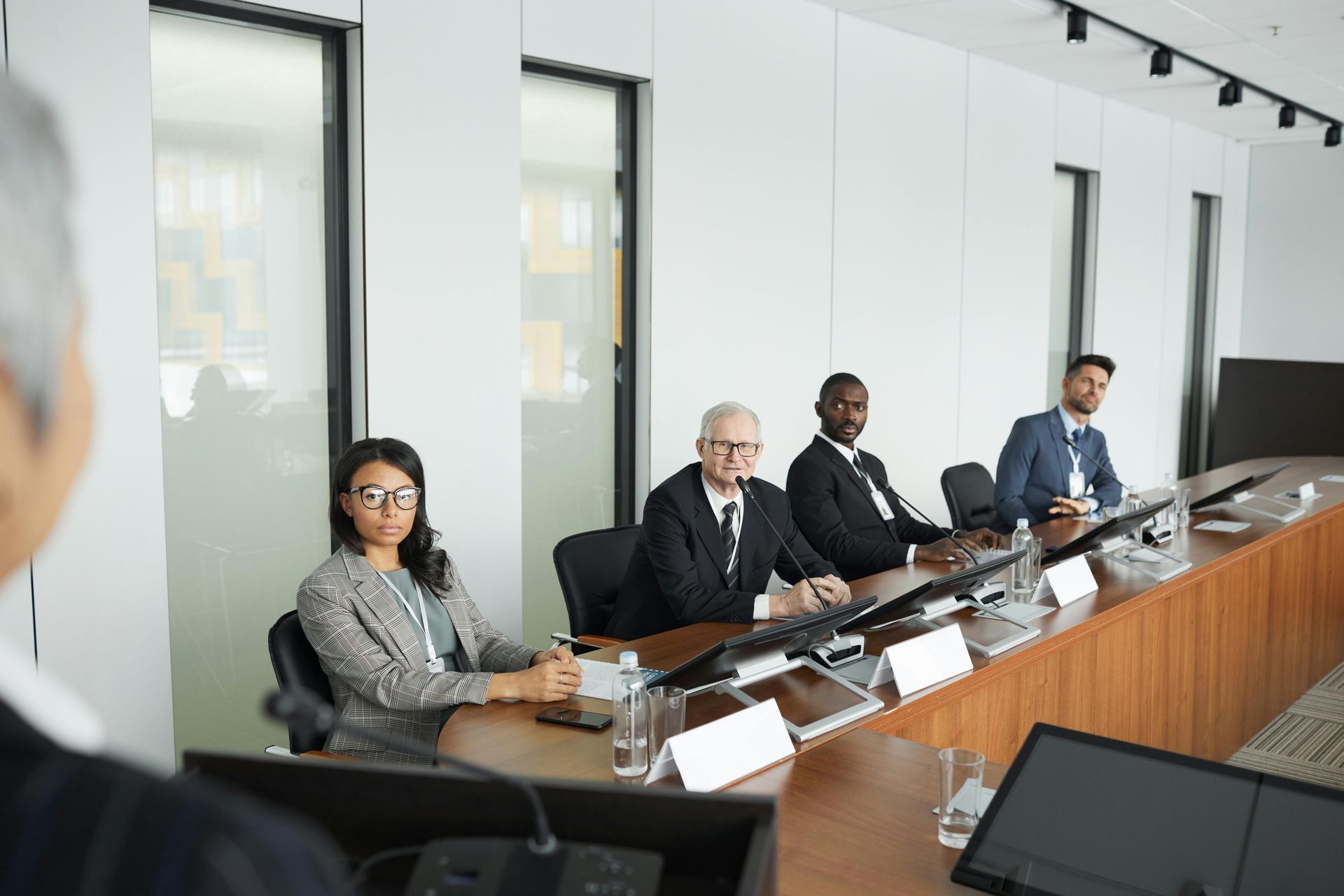A group of people are sitting around a table in a conference room while a man writes on a glass wall.
