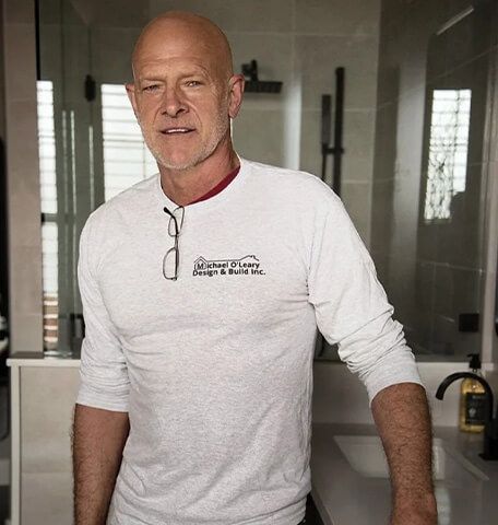 A bald man wearing a white shirt and glasses is standing in a bathroom.