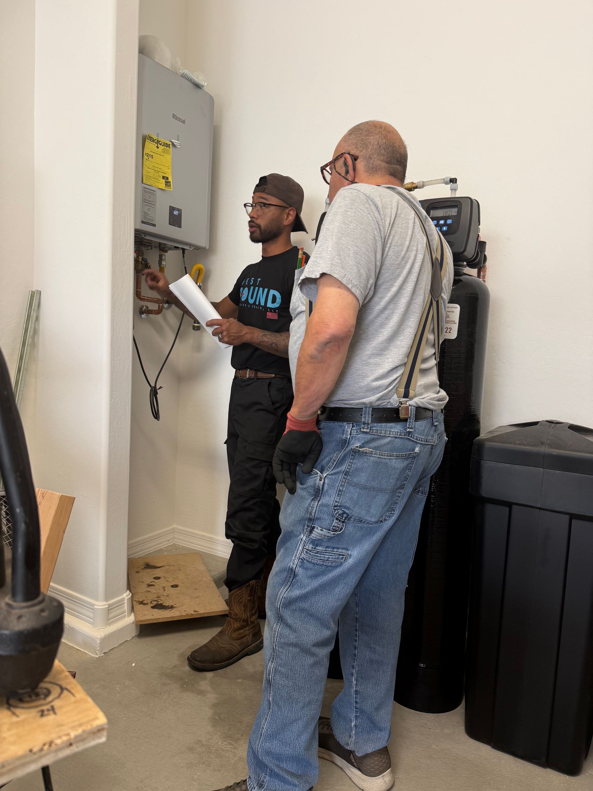 Two people, one holding papers, looking at a utility panel. Indoors, next to a water softener.