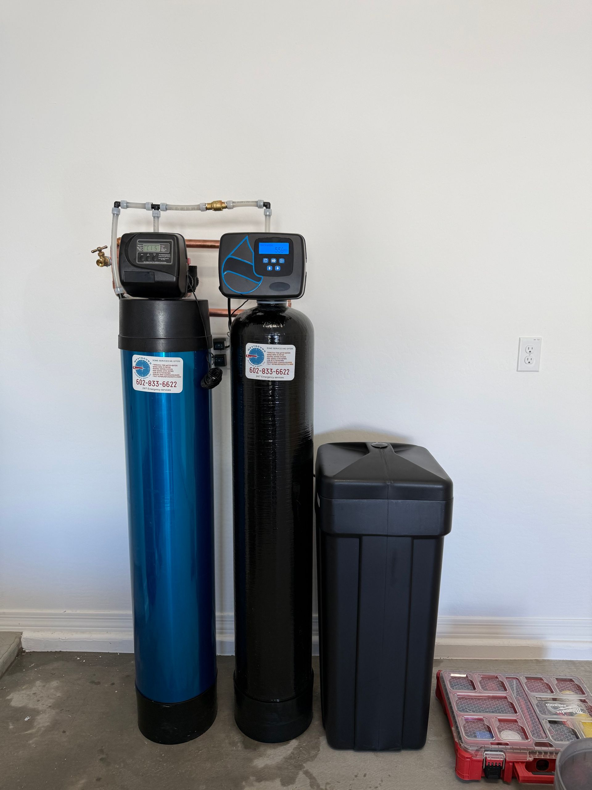 Water filtration system: two tall tanks, blue and black, with digital control panels, and a black brine tank, against a white wall.