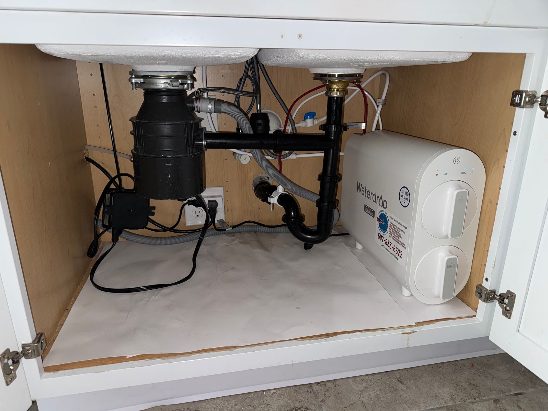 Under-sink view: garbage disposal, plumbing, and a water filtration system within a white cabinet.