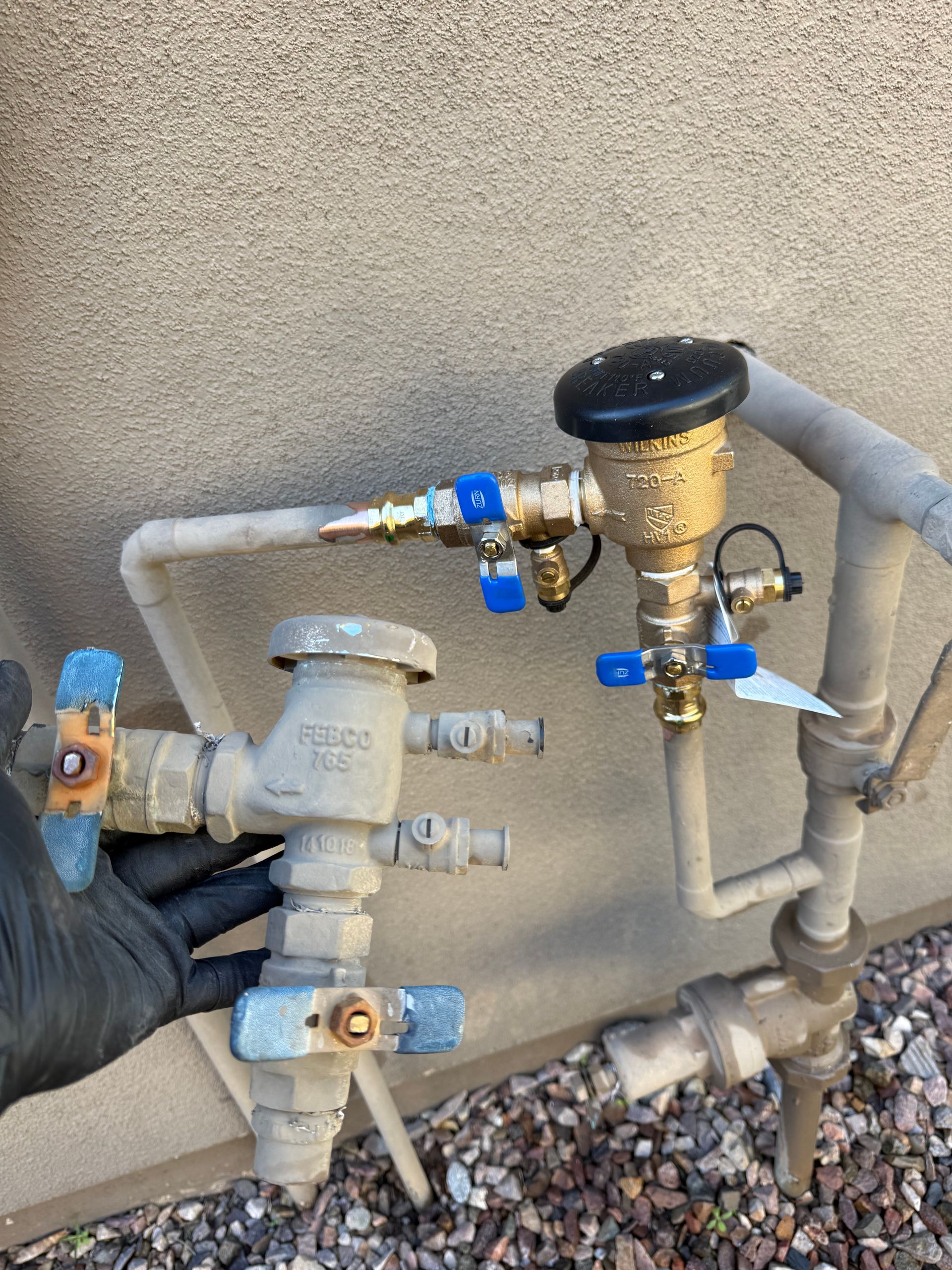Hand holding a water valve next to exterior plumbing components. Includes metal valves, pipes, and a black glove.