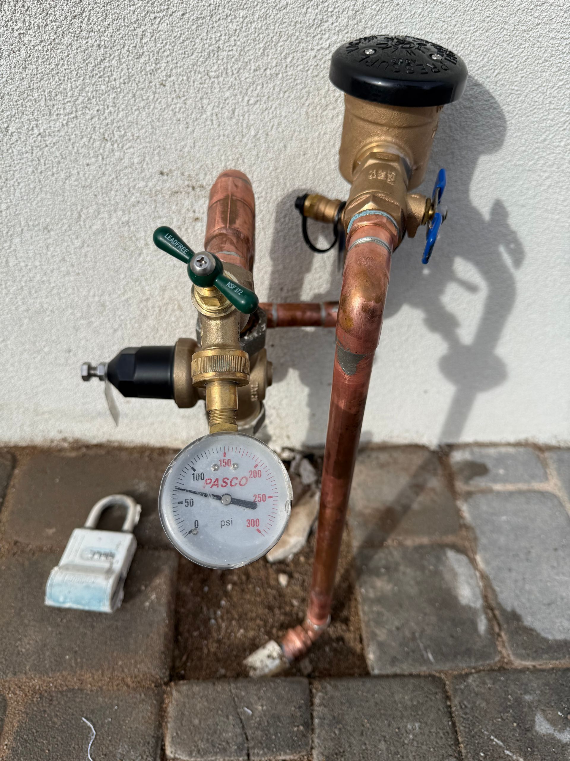 Copper pipes, water pressure gauge, and irrigation control valves against a stucco wall. A padlock is near the gauge.