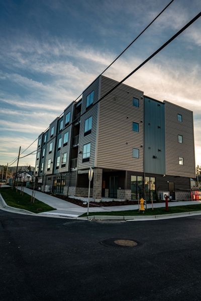 Modern apartment building at a street corner with blue and grey facade, power lines overhead, sunset.