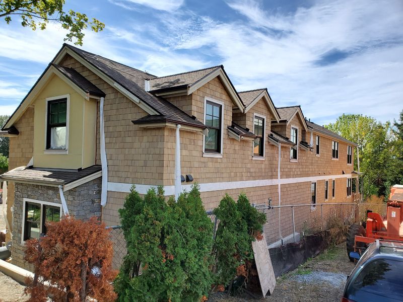 Tan shingled building with multiple dormers, dark windows, and brown trim under a blue sky.
