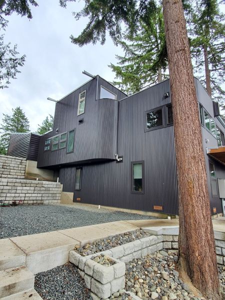 Modern black house with angled walls and windows, set in a wooded area with stone retaining walls.