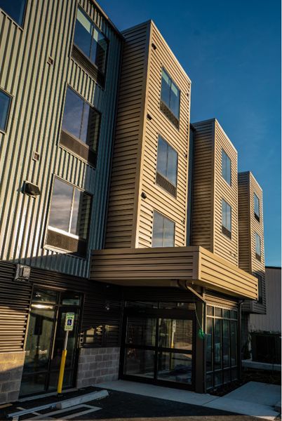 Modern multi-story building exterior with vertical corrugated metal siding, entry canopy, blue sky.