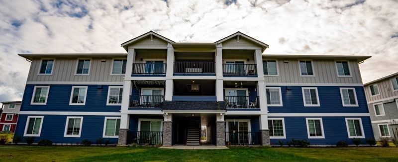 Multi-story blue and gray apartment building with balconies, set against a cloudy sky.