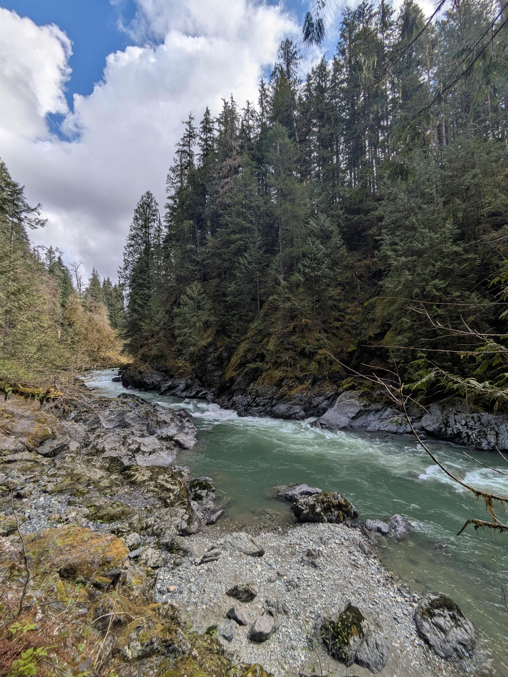 River flowing through a rocky, forested landscape under a cloudy blue sky.