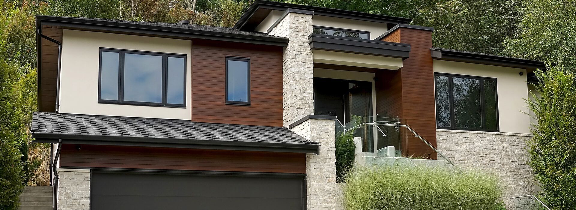 Modern home with cream siding, brown wood accents, and a dark gray garage door, nestled in lush greenery.