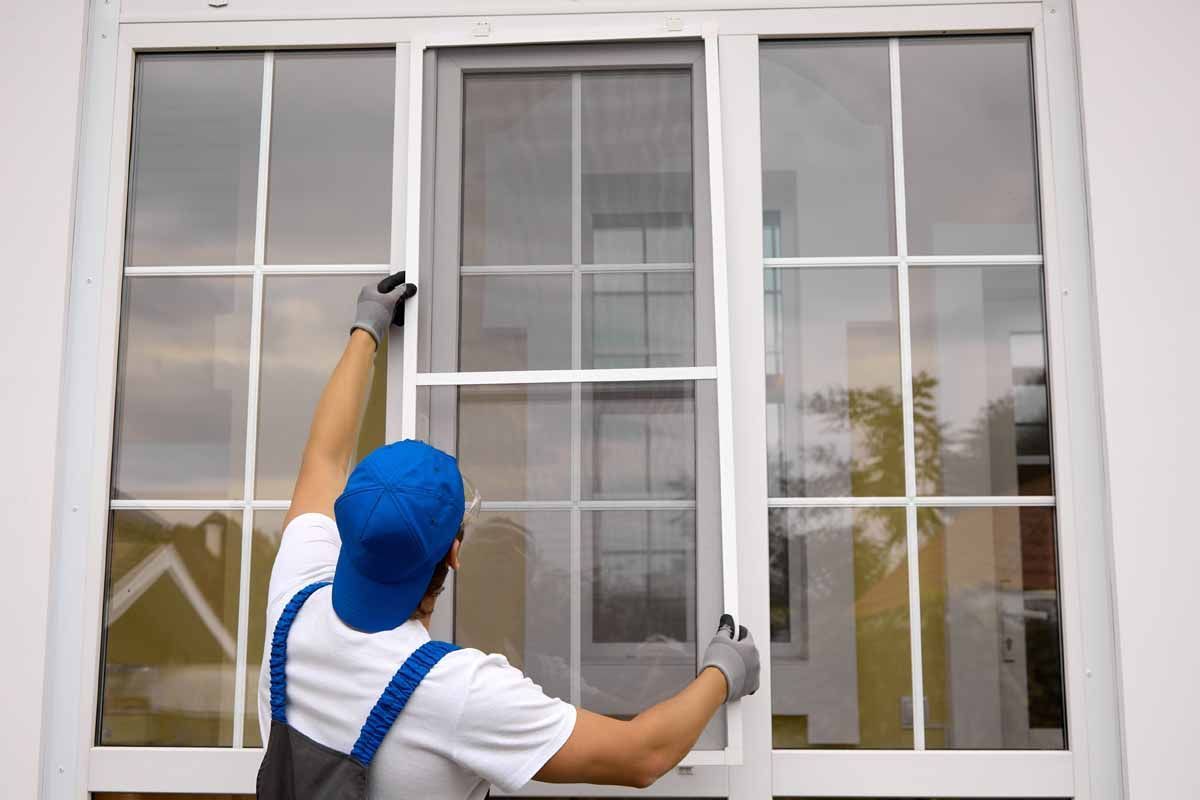 Man in blue cap and work gloves installing a window screen on a white building.
