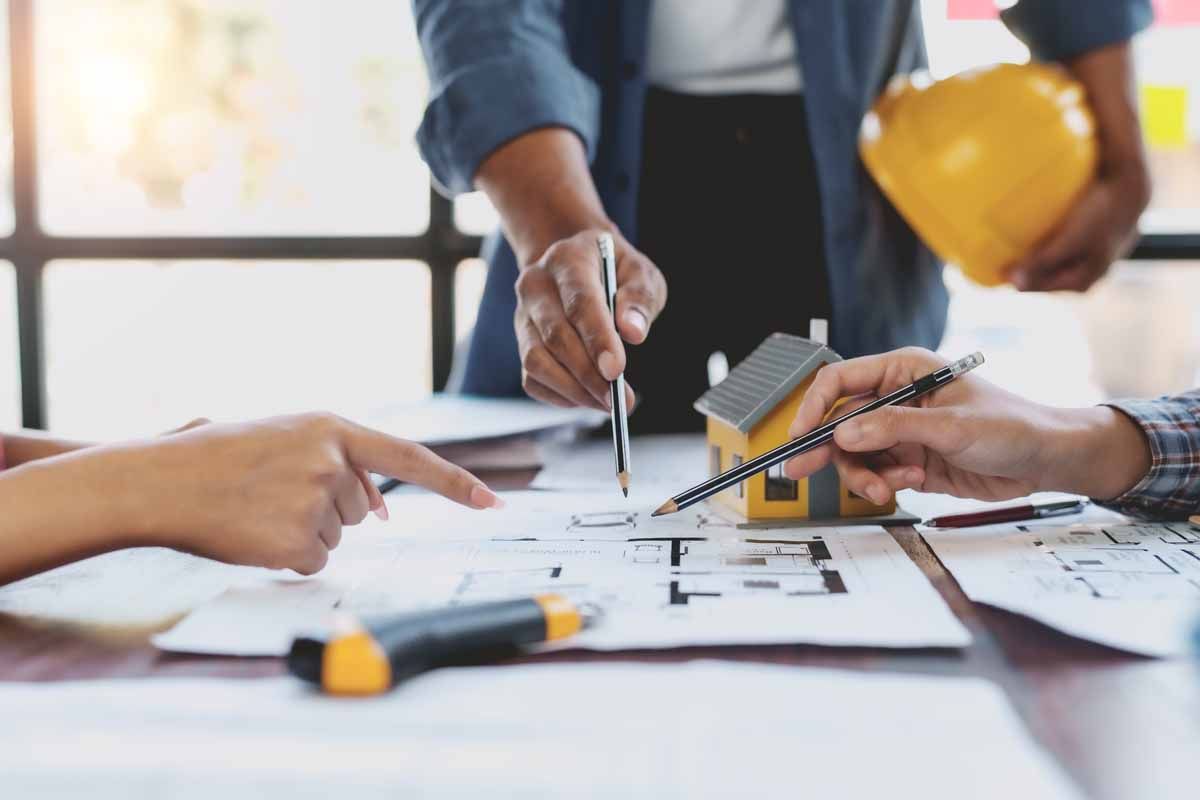 Architects reviewing blueprints, pointing, discussing plans at a table with a miniature house and hardhat.