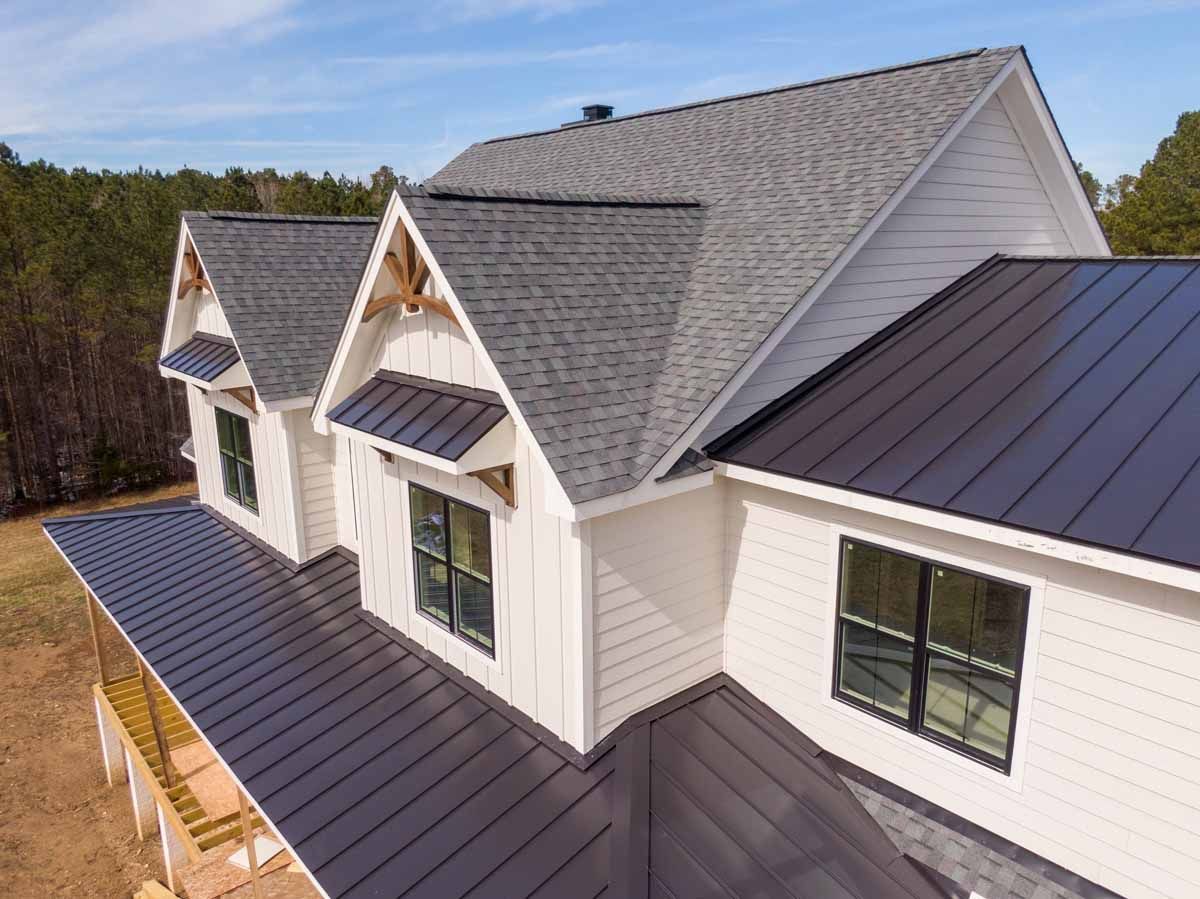 Modern white house with gray and black metal roofing, dormers, and forest backdrop.