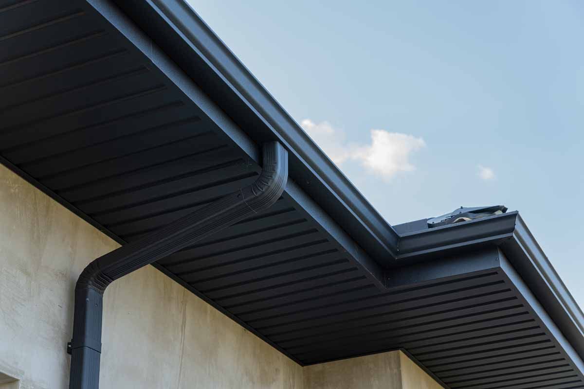 Dark gray rain gutter system on a beige building, with a blue sky background.