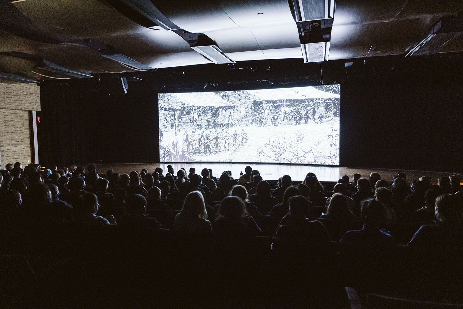 A crowd of people are sitting in front of a large screen in a dark room.