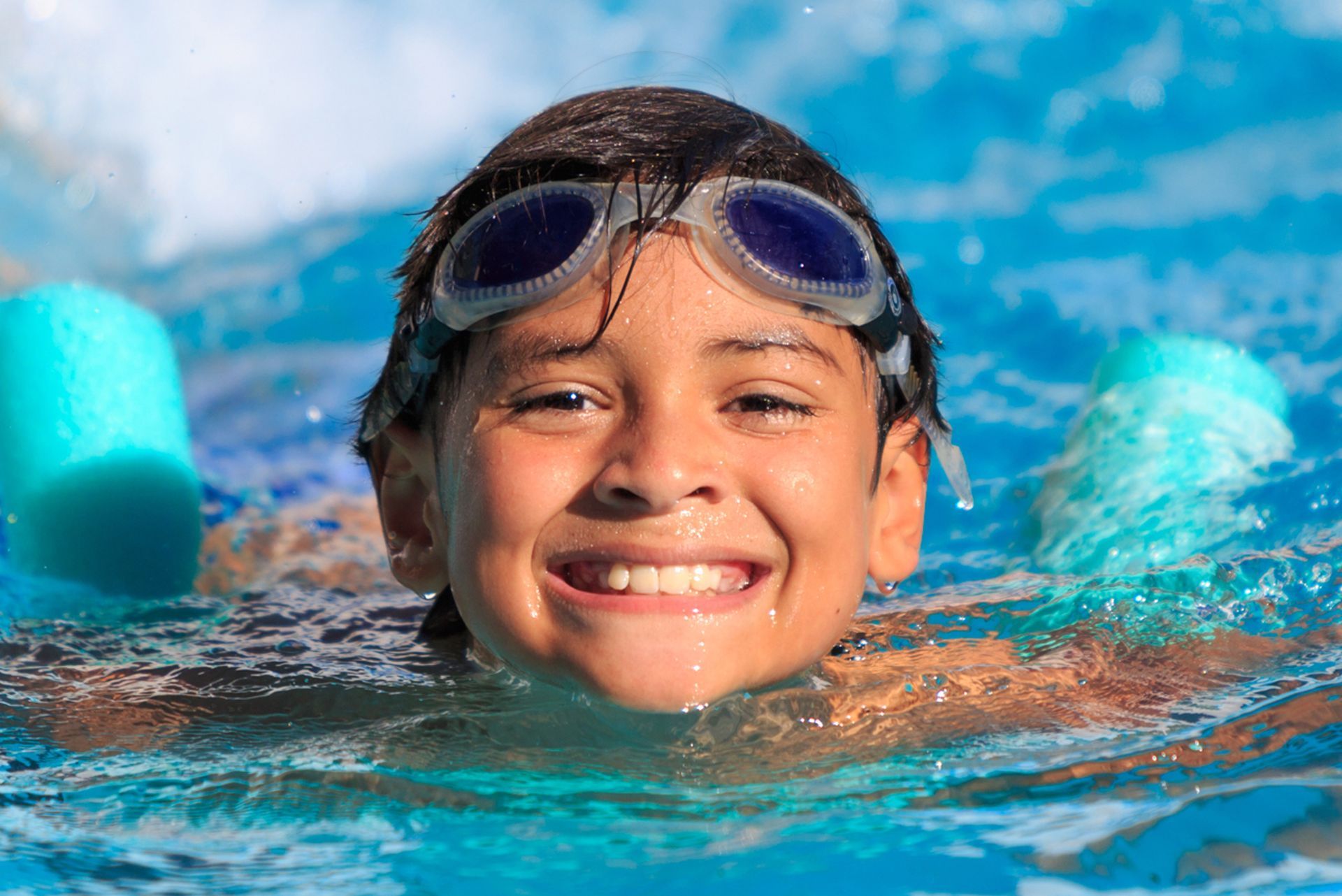 A young boy wearing goggles is swimming in a pool and smiling.