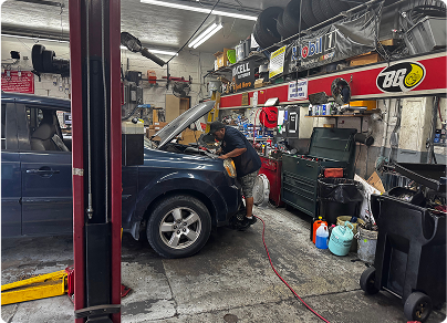 Mechanic working on a blue car in a cluttered auto repair shop. Tools, tires, and a lifted car.