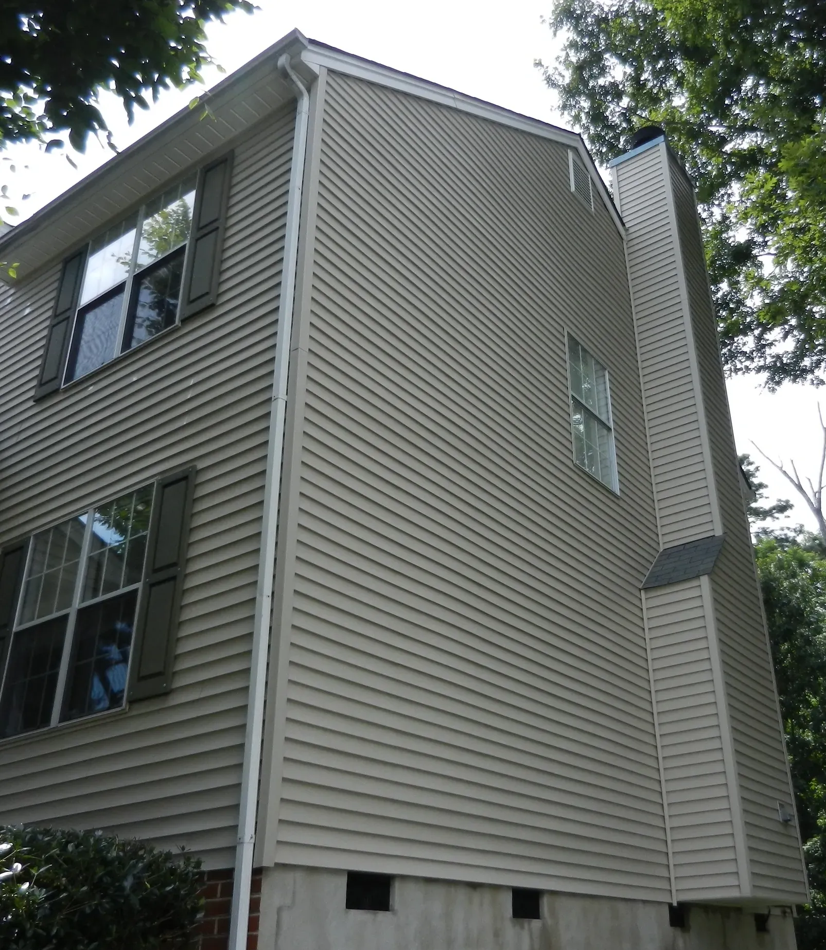 Side of a two-story beige house with a tall chimney, green shutters, and windows; surrounded by trees.