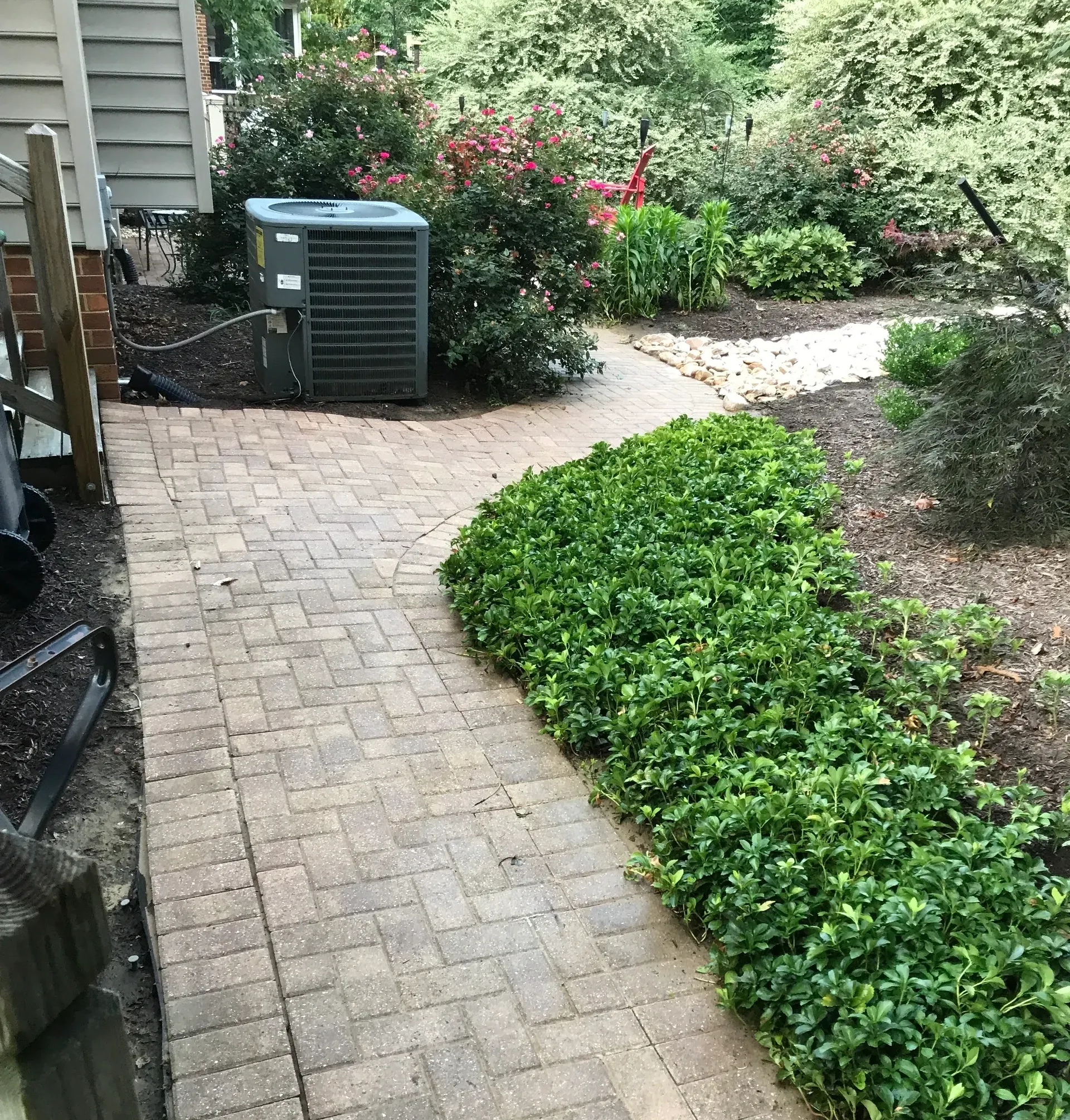 Brick pathway curves through a garden, past an air conditioner unit and green plants.