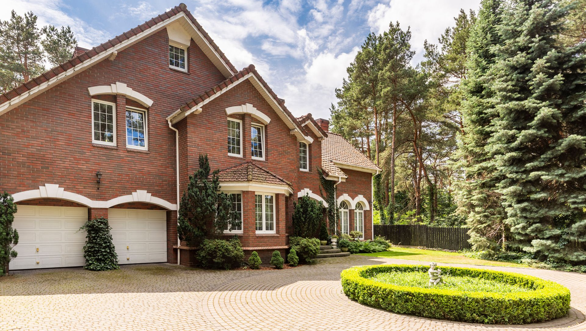 Brick house with white garage doors, curved driveway, and manicured landscaping under a cloudy sky.