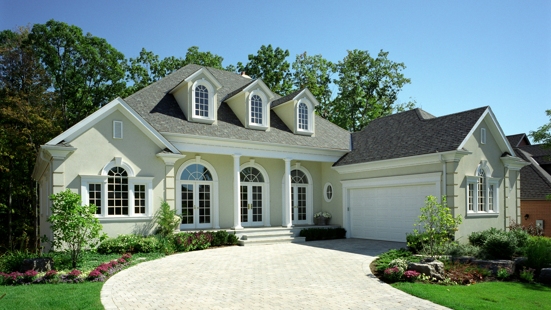 Cream-colored house with columns, arched windows, and a circular driveway. Lush green landscaping and blue sky.