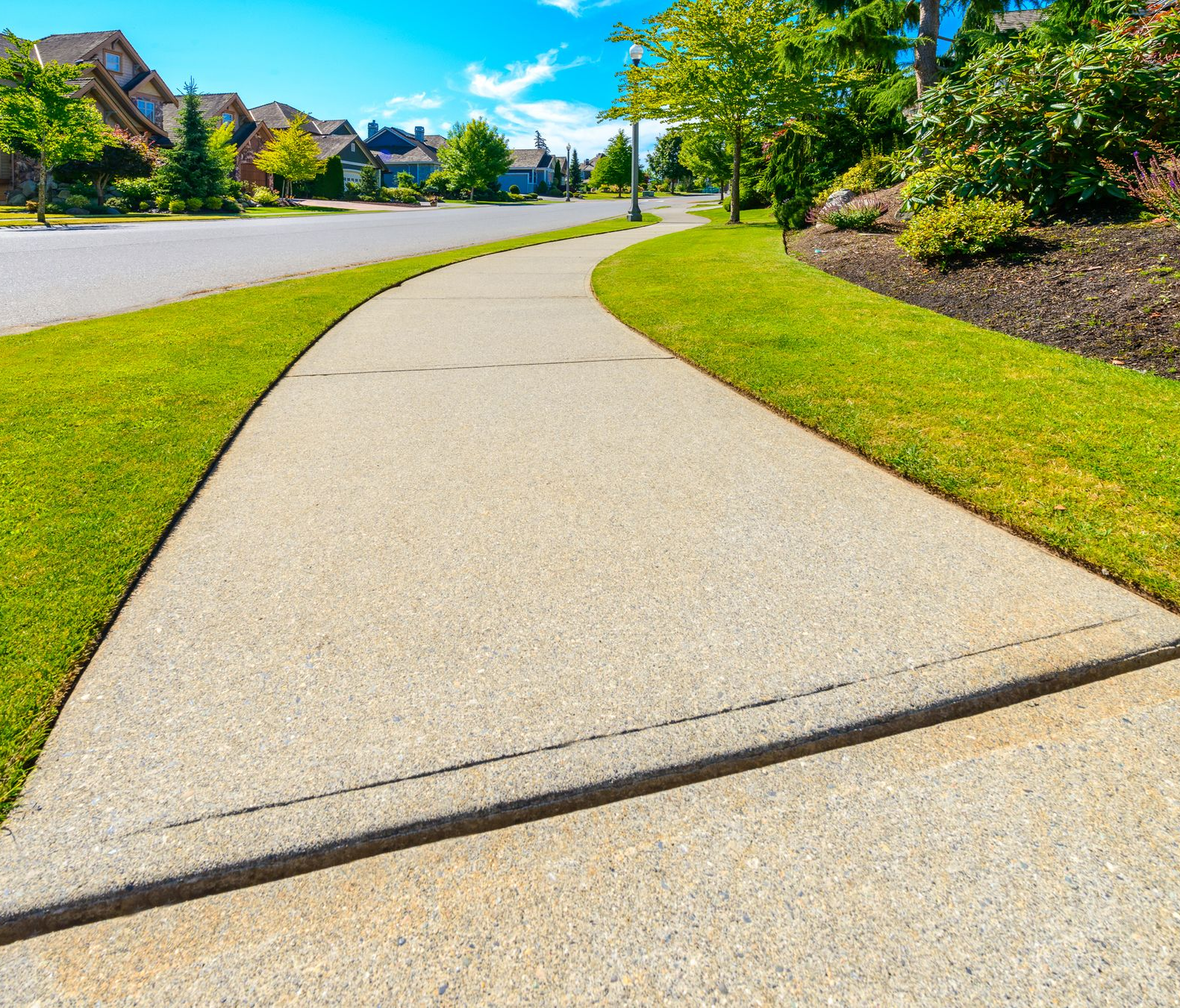 Concrete sidewalk curves through a neighborhood with green grass, houses, and blue sky.
