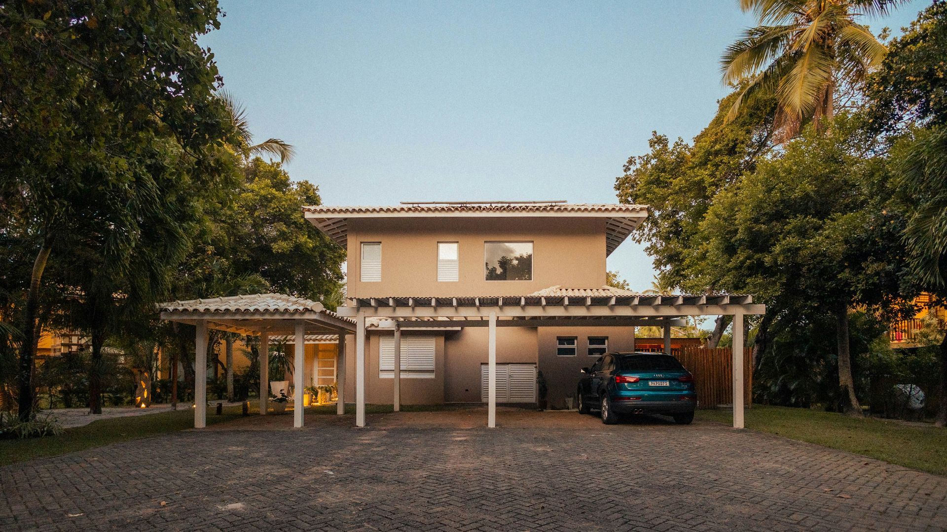 Two-story beige house with a covered carport, a dark blue car, and surrounded by trees.