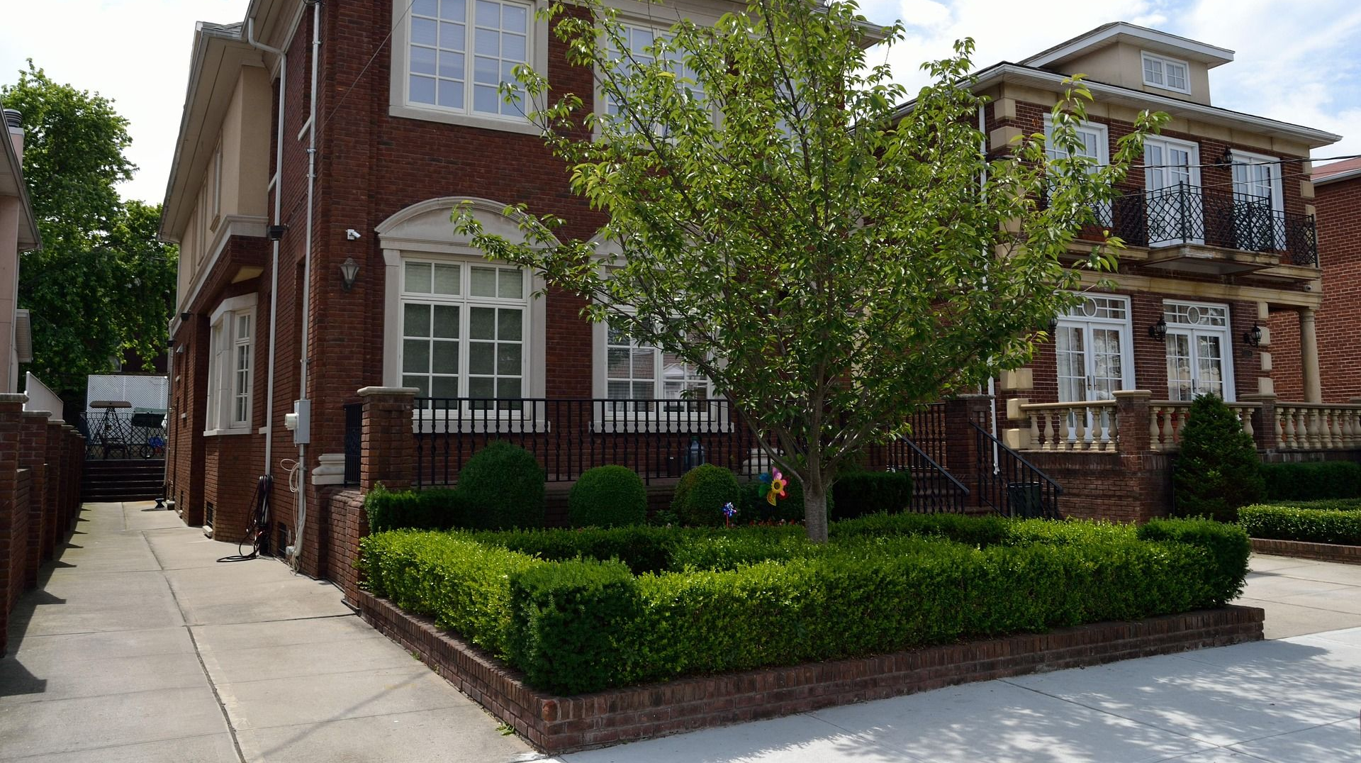 Two brick houses with manicured hedges and a central tree under a blue sky with white clouds.