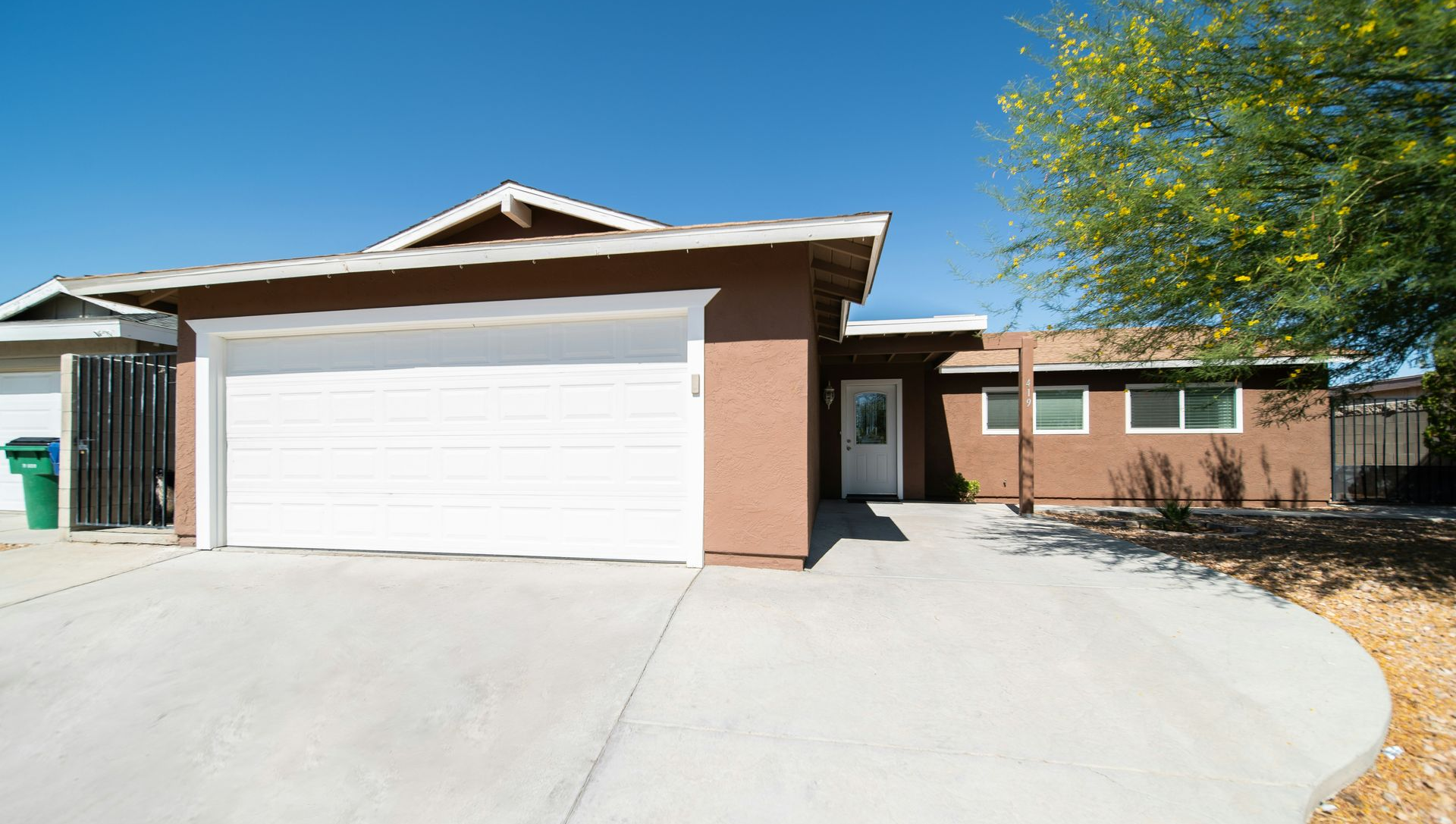 A single-story brown house with a large white garage door, concrete driveway, and a yellow-flowering tree under blue sky.