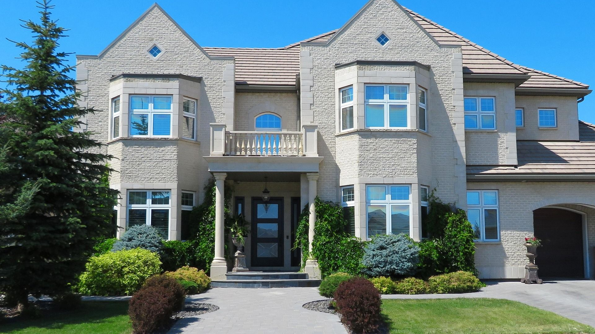 Beige two-story house with bay windows, balcony, and arched entryway under a clear blue sky.