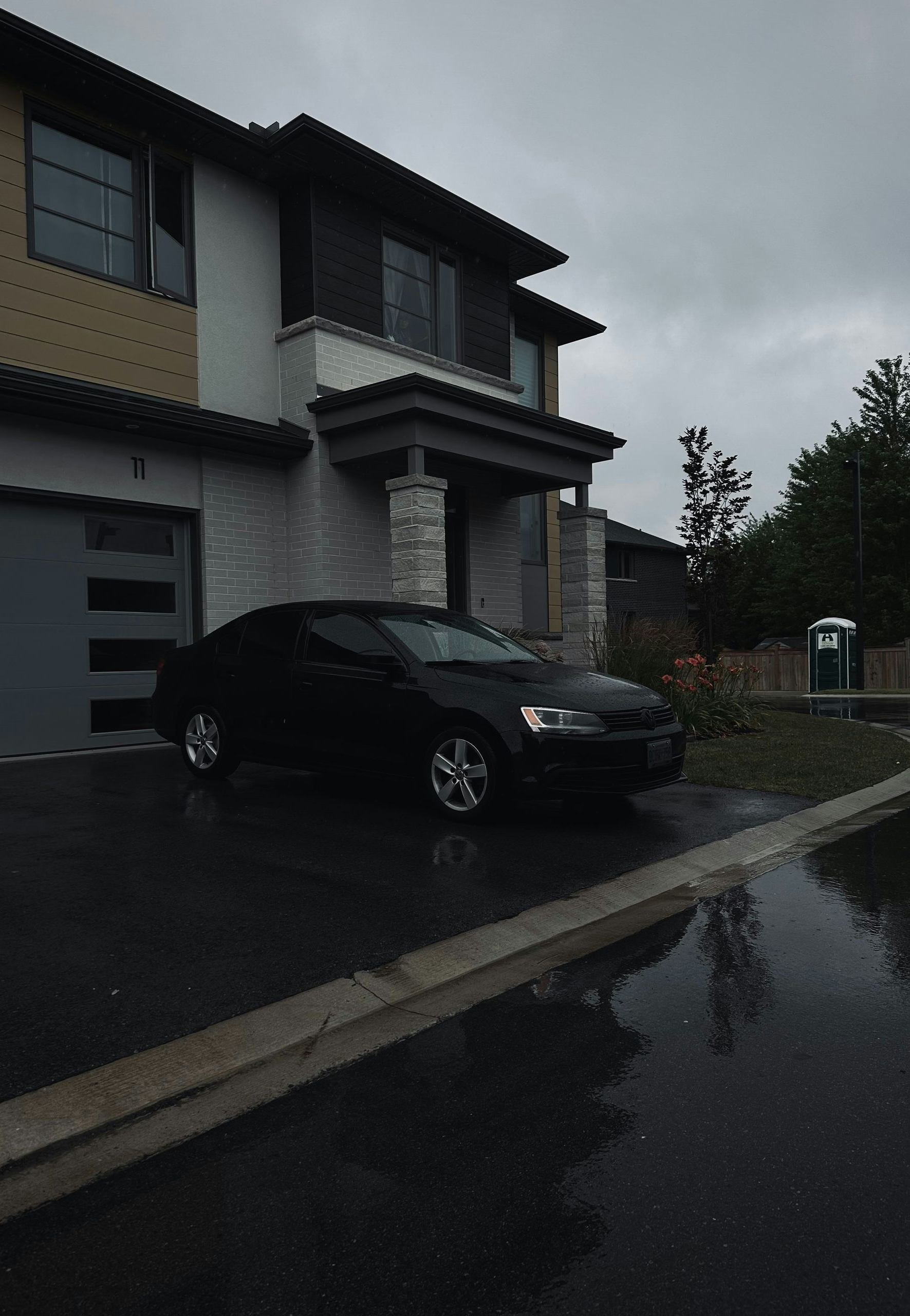 Black car parked in front of a two-story house with a garage, on a wet, paved road under a cloudy sky.