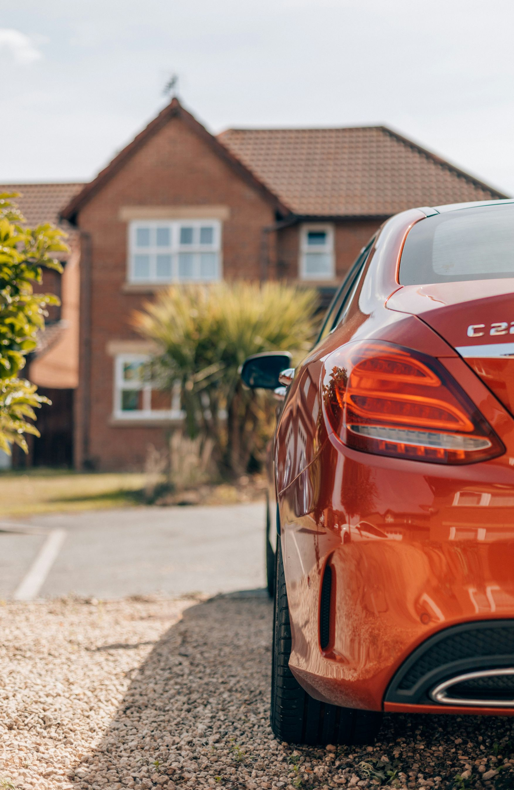 Orange Mercedes-Benz C220 parked on gravel in front of a brick house on a sunny day.