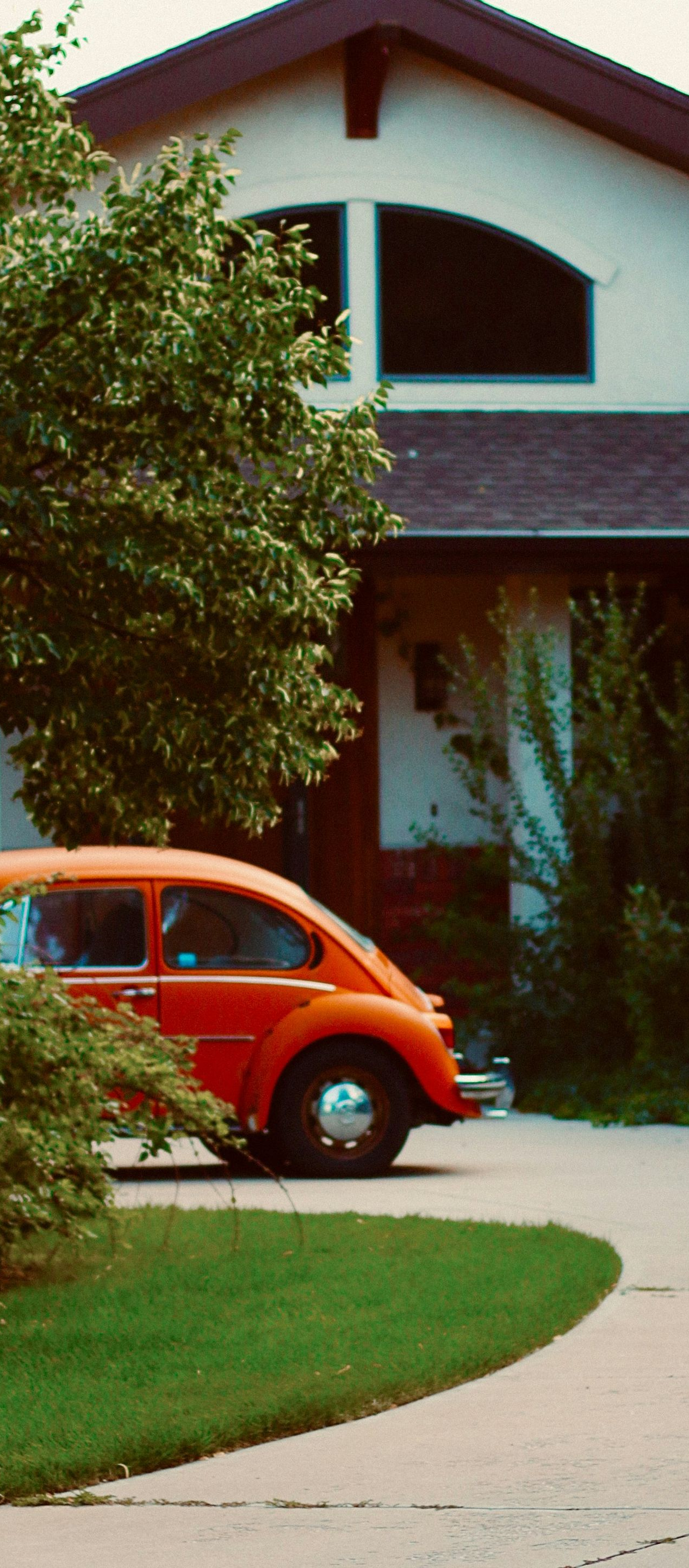 A bright orange Volkswagen Beetle parked on a driveway in front of a house with an arched window.
