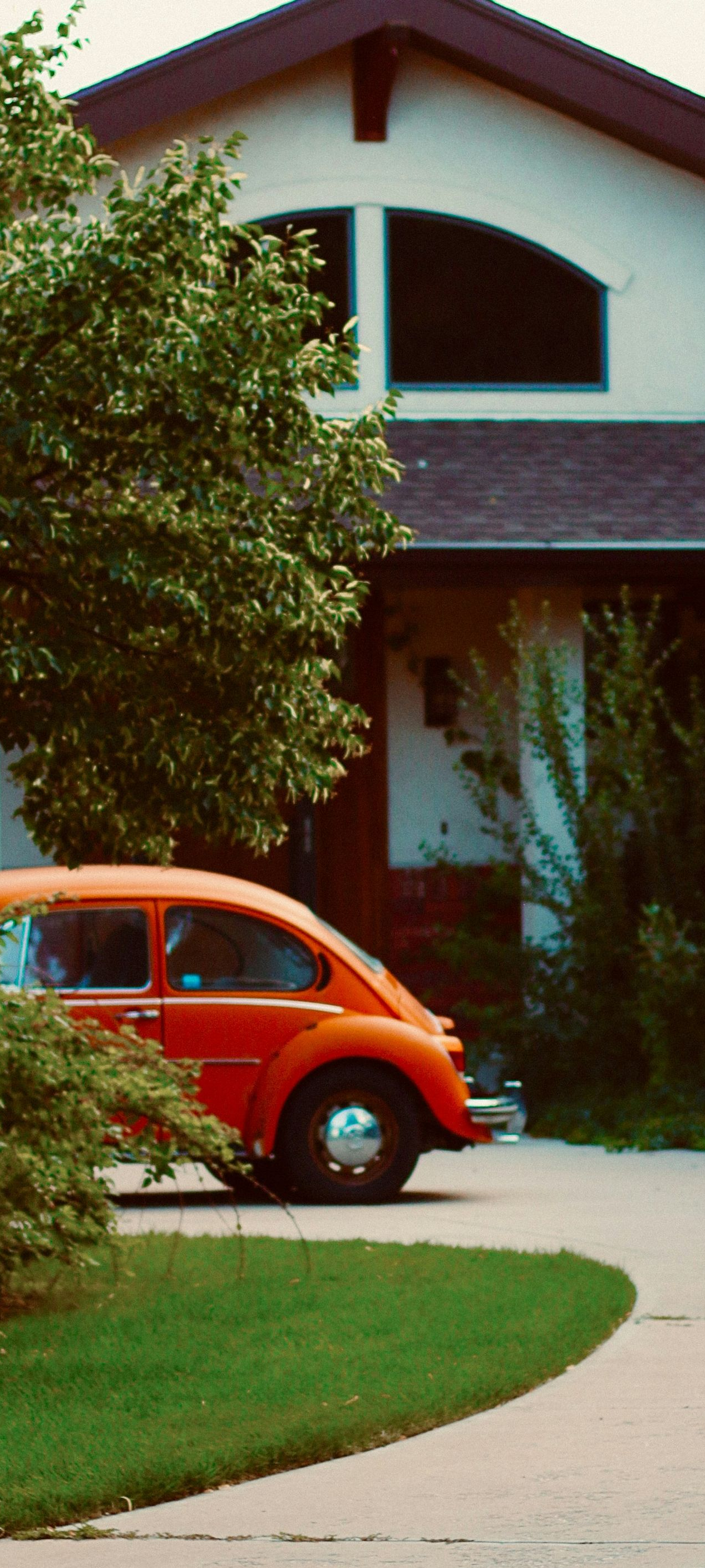 A bright orange Volkswagen Beetle parked on a driveway in front of a house with an arched window.