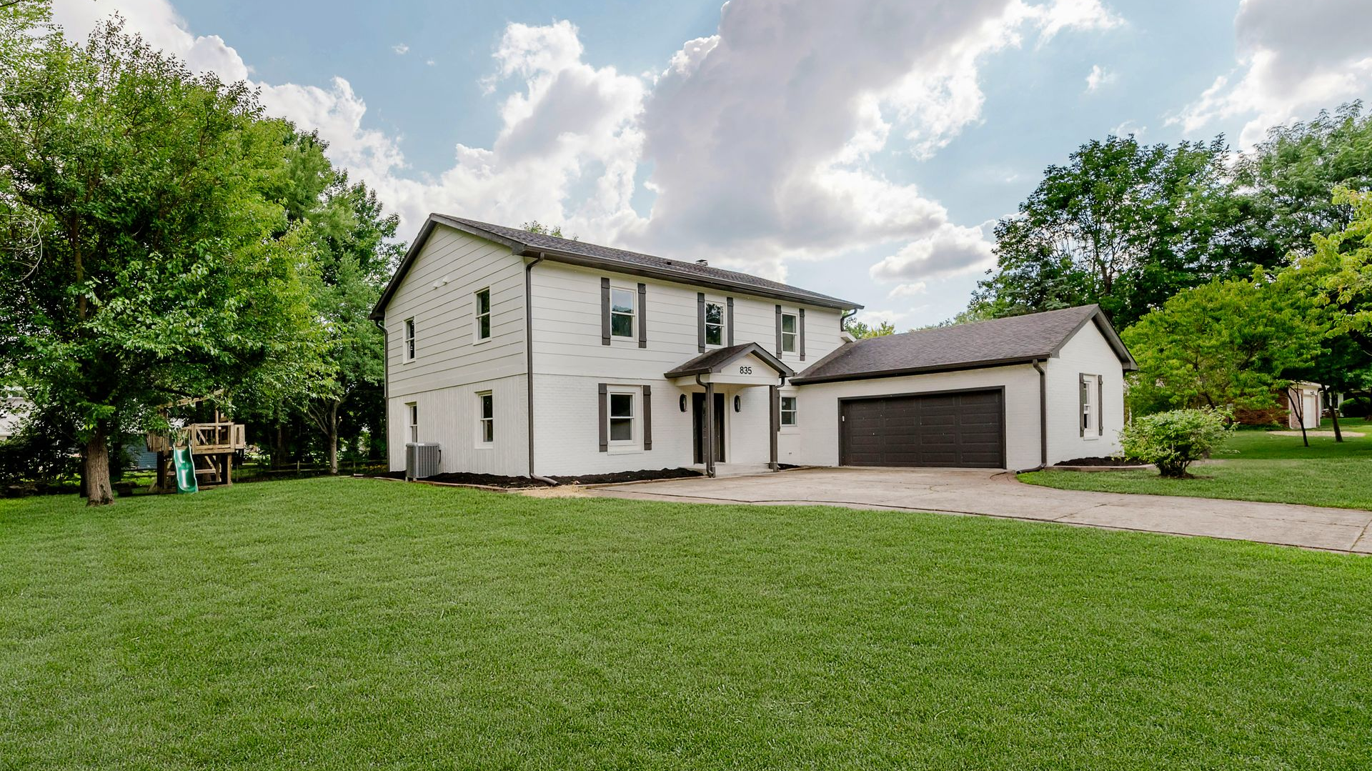 A white two-story house with dark shutters and a matching garage sits on a lush green lawn under a bright, cloudy sky.